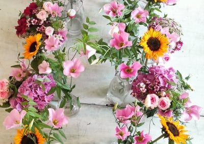 Floristik für Hochzeit in Dresden: Mehrere kleine Glasvasen mit bunten Sommerblumen rosa Rosen und Sonnenblumen als Tischschmuck.